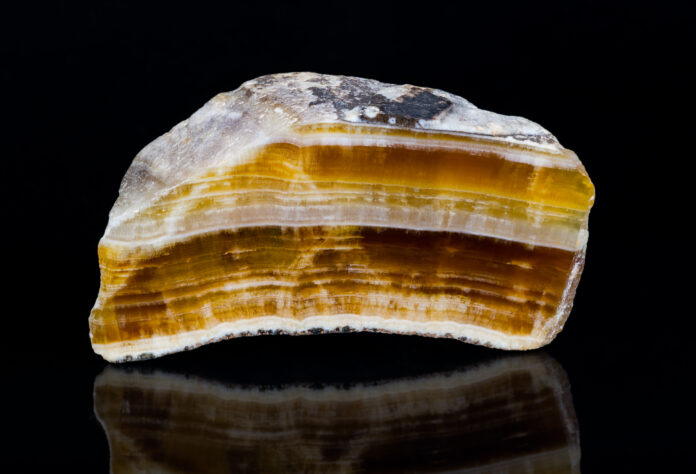 Cut polished aragonite gemstone with reflection on black background. Close-up of beautiful yellow, brown and white striped cross-section of mineral from Hridelec near Nova Paka in Czechia. Mineralogy. Cut polished aragonite gemstone with reflection on black background. Close-up of beautiful yellow, brown and white striped cross-section of mineral from Hridelec near Nova Paka in Czechia. Mineralogy.