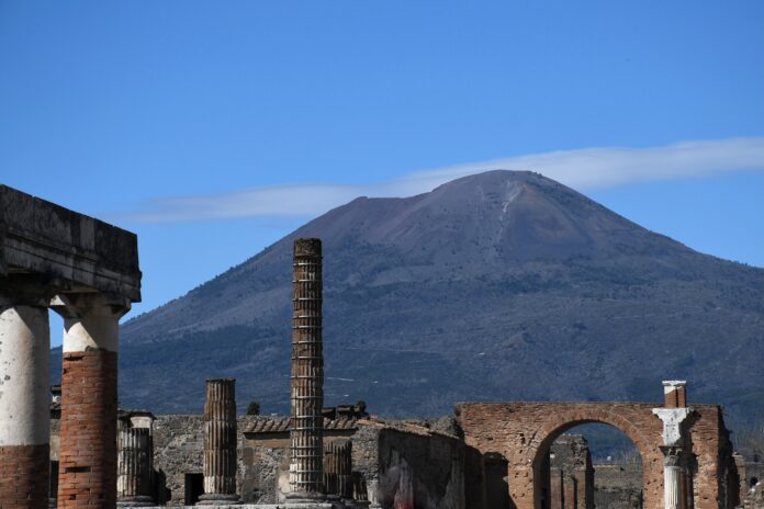Mt Vesuvius rises up above the ruins of the Forum in Pompeii. A road can be seen zig-zagging across the mountain. The sky is blue. Mt Vesuvius rises up above the ruins of the Firum in Pompeii.