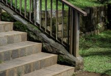 Landscaping With Rocks: Ideas for Using Stone Outdoors Stone stairs with a metal handrail leading through a lush garden. The scene features a mossy stone retaining wall and green grass, highlighting peaceful outdoor landscaping with rocks