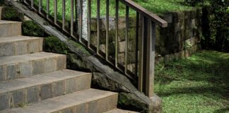 Stone stairs with a metal handrail leading through a lush garden. The scene features a mossy stone retaining wall and green grass, highlighting peaceful outdoor landscaping with rocks