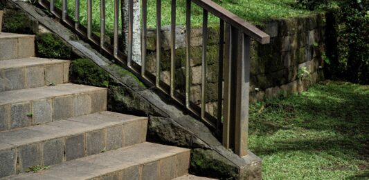 Landscaping With Rocks: Ideas for Using Stone Outdoors Stone stairs with a metal handrail leading through a lush garden. The scene features a mossy stone retaining wall and green grass, highlighting peaceful outdoor landscaping with rocks