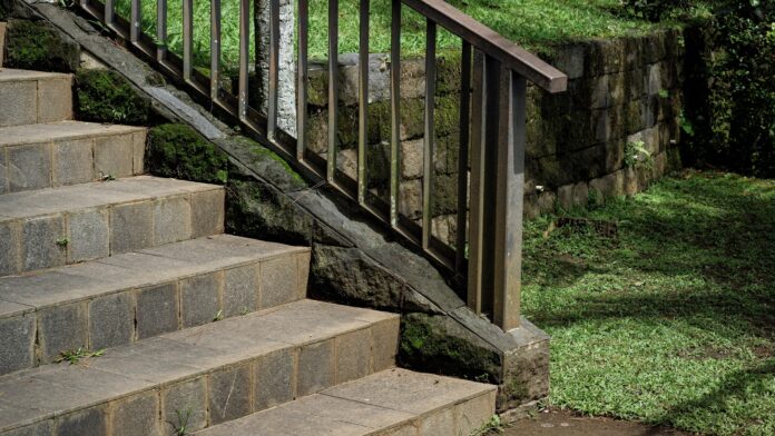 Landscaping With Rocks Stone stairs with a metal handrail leading through a lush garden. The scene features a mossy stone retaining wall and green grass, highlighting peaceful outdoor landscaping with rocks