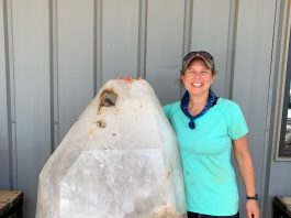 rockhounding woman standing next to giant crystal as tall as her